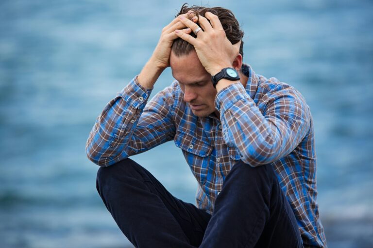 Un homme en chemise à carreaux est assis au bord de l'eau, l'air en détresse, symbolisant le stress.